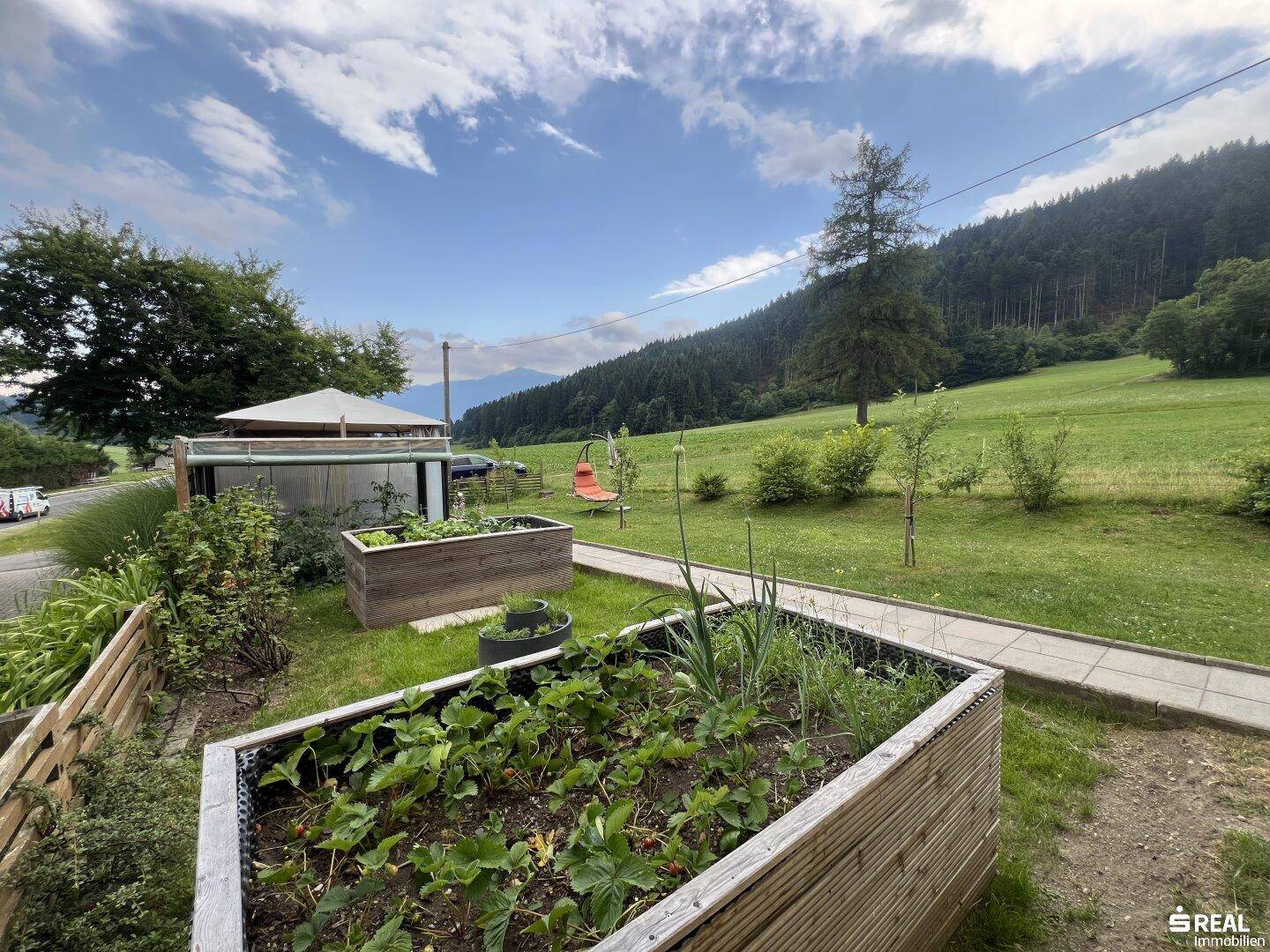 Gepflegter Garten mit Hochbeeten und Schaukelstuhl, umgeben von grünen Wiesen und Bergen.
