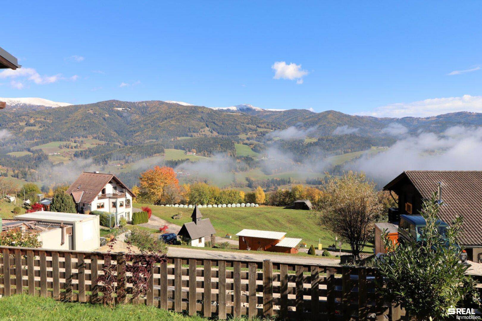 Panoramablick über ein malerisches Tal mit herbstlicher Vegetation und nebelverhangenen Bergen im Hintergrund.