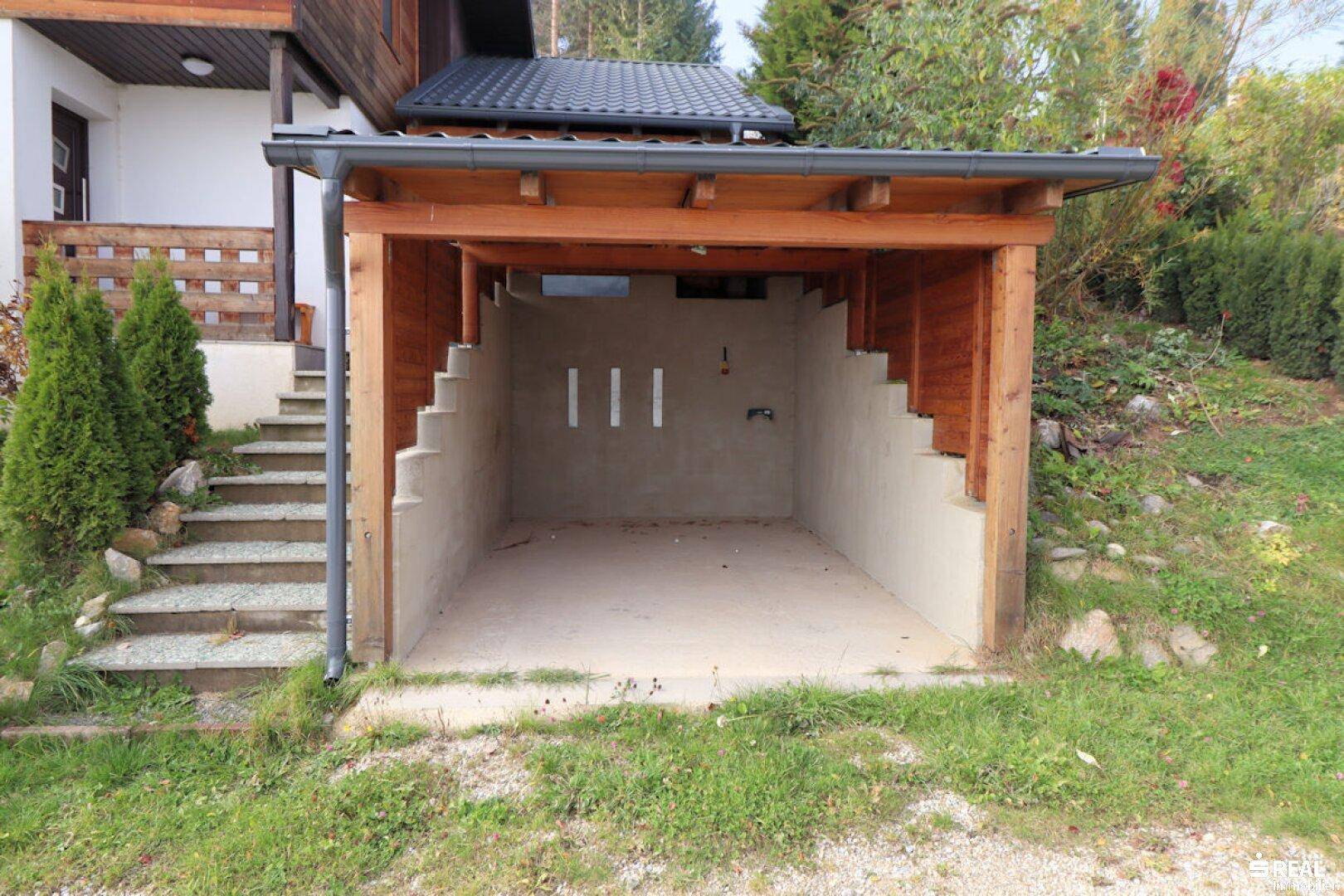 Ein offener Holzcarport bietet einen überdachten Stellplatz, integriert in die natürliche Umgebung des Hauses.