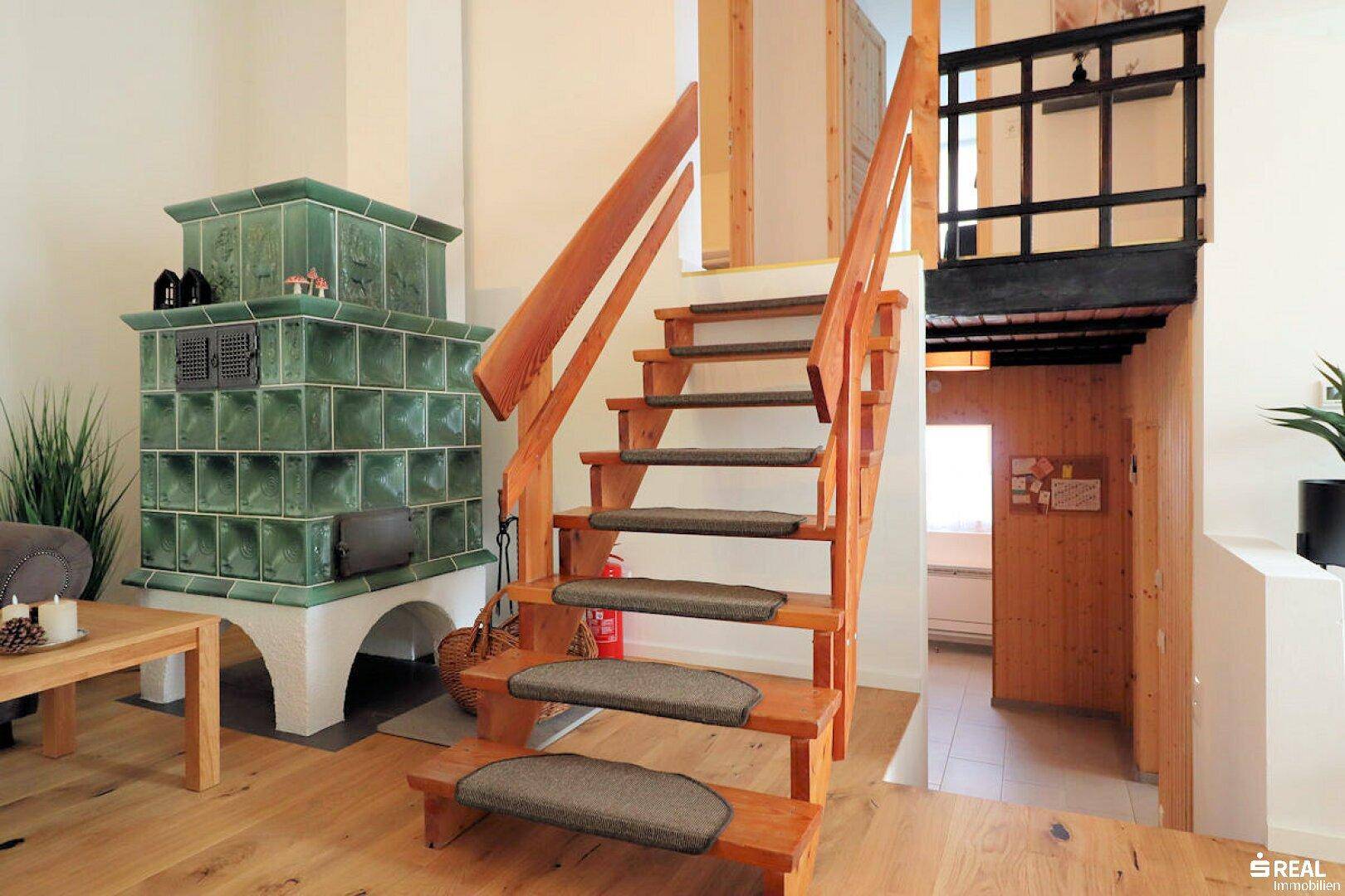 Heller Wohnbereich mit einer Holztreppe, die in die obere Etage führt, und einem markanten Kachelofen.