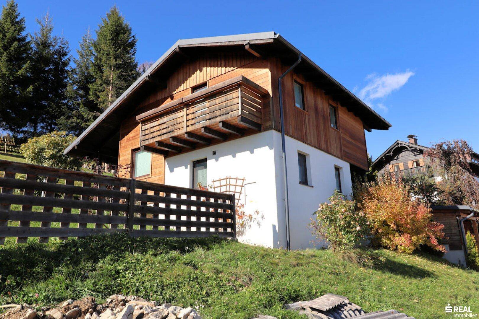 Das Holzchalet steht auf einem grünen Hügel, umgeben von einem Holzzaun und Bäumen unter blauem Himmel.