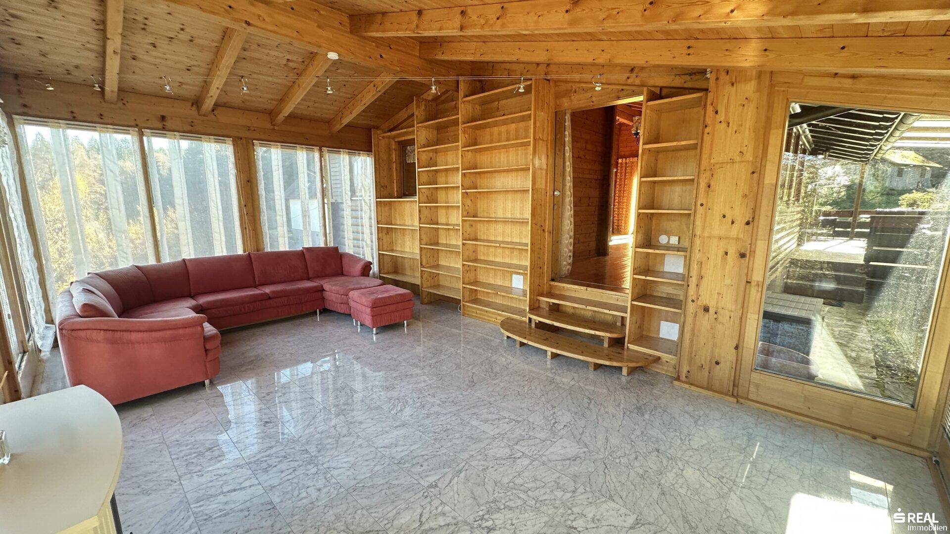 Heller Wintergarten mit rotem Sofa, Fliesenboden und großen Fenstern mit Blick ins Grüne.