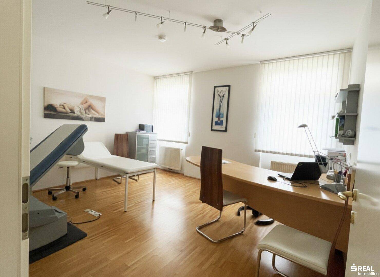 Helles Behandlungszimmer mit Untersuchungsliege, Schreibtisch und großen Fenstern, ausgestattet für medizinische Praxis.