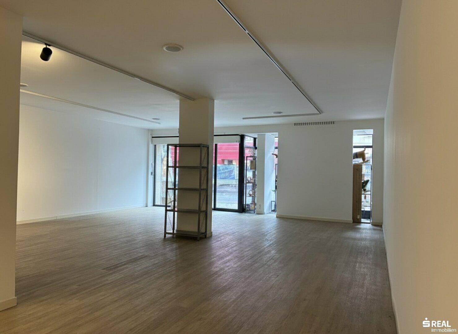 Offener Gewerberaum mit großen Schaufenstern zur Straße und Holzboden, ideal für Einzelhandel oder Ausstellungen.