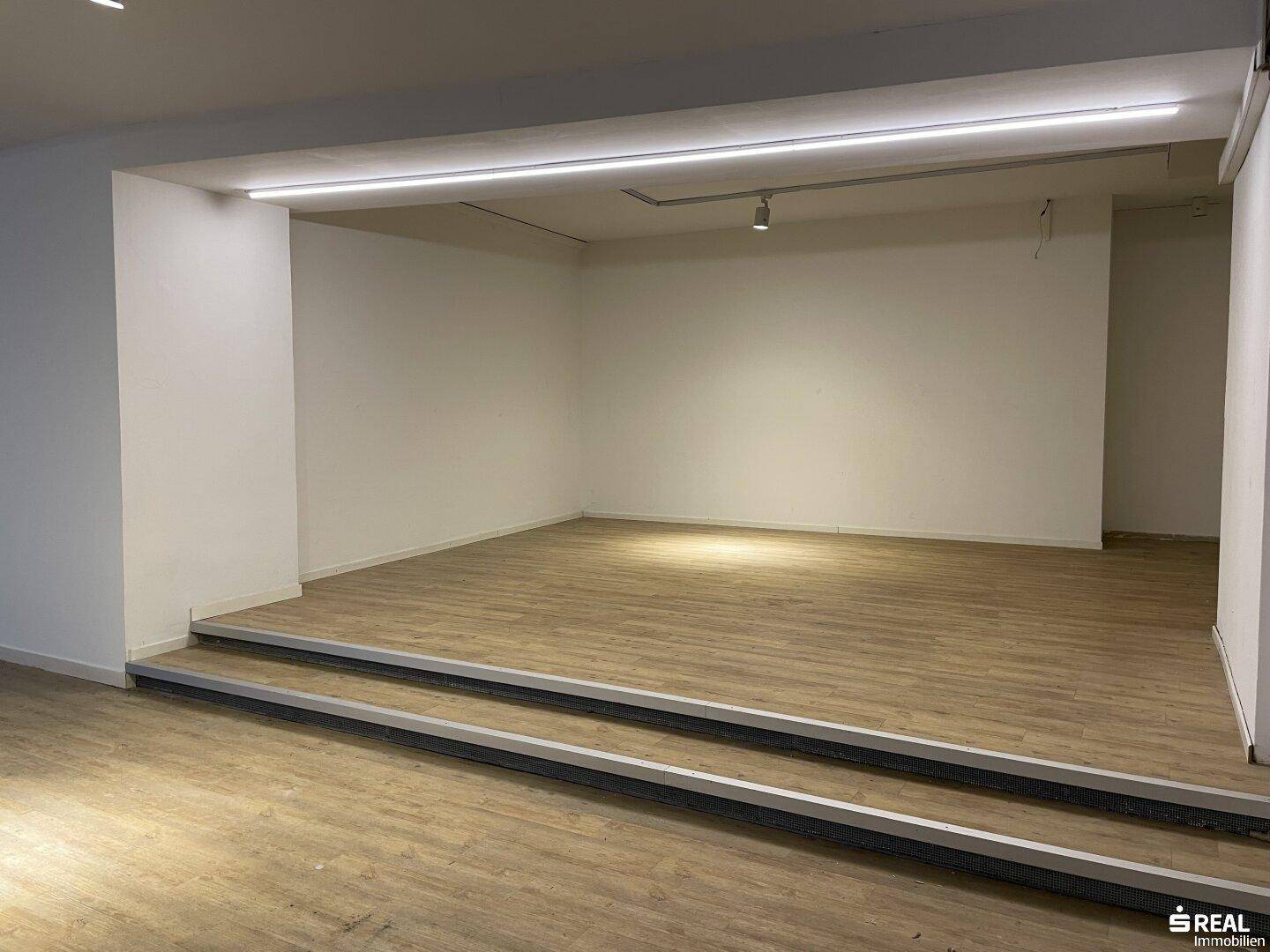 Erhöhter Bereich in einem Gewerberaum mit Holzboden und indirekter Beleuchtung, vielseitig nutzbar für Präsentationen.