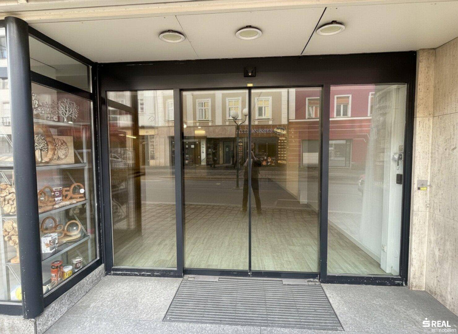 Eingangsbereich eines Geschäfts mit großen Glastüren und Schaufenstern, reflektiert die städtische Umgebung.