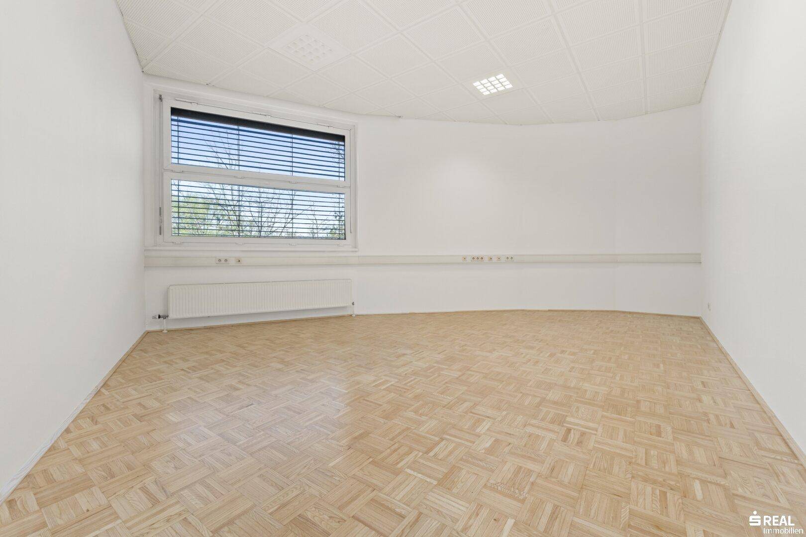 Heller, leerer Büroraum mit Parkettboden und einem großen Fenster mit Jalousien.