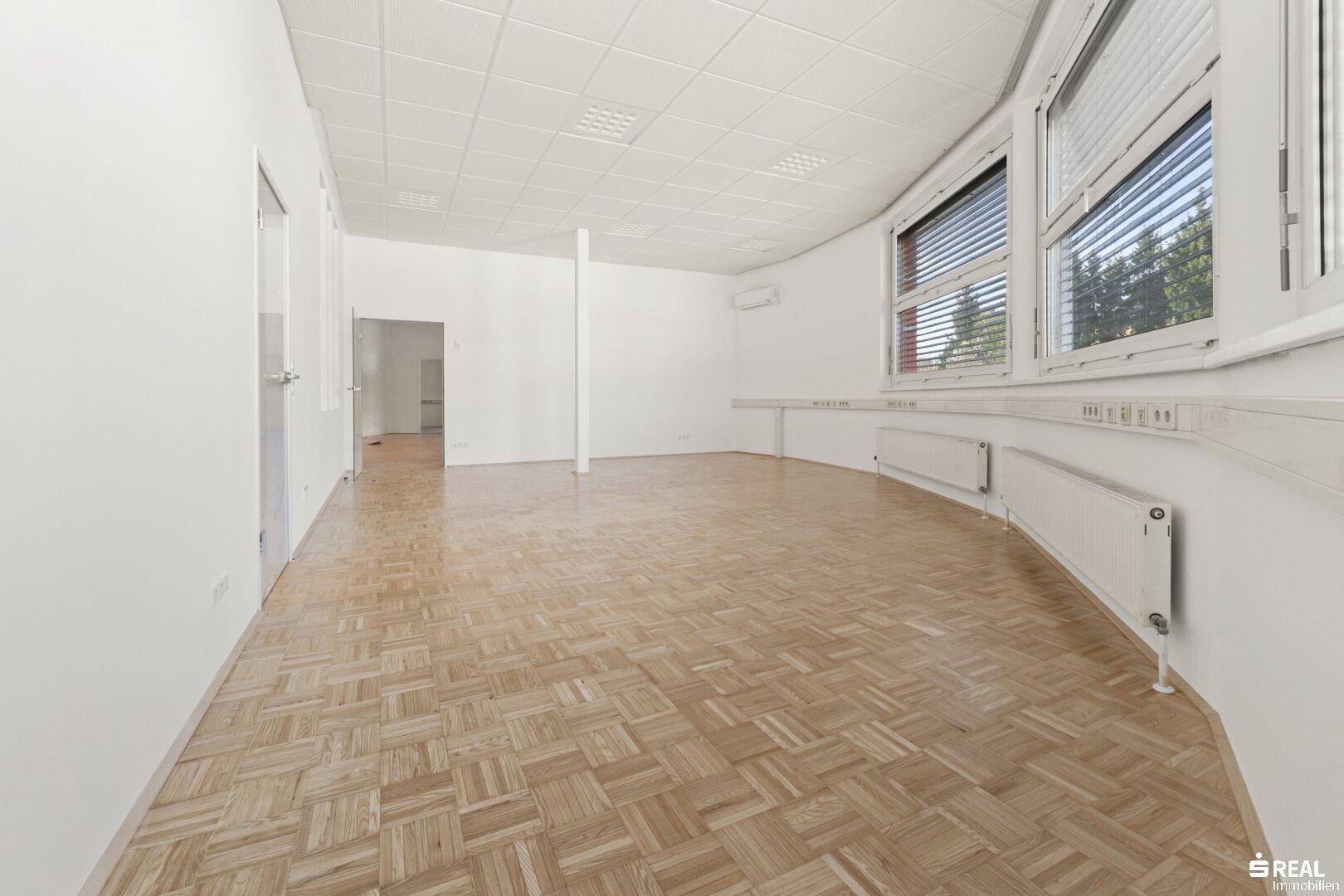 Großer, leerer Büroraum mit Parkettboden, weißen Wänden und mehreren Fenstern.