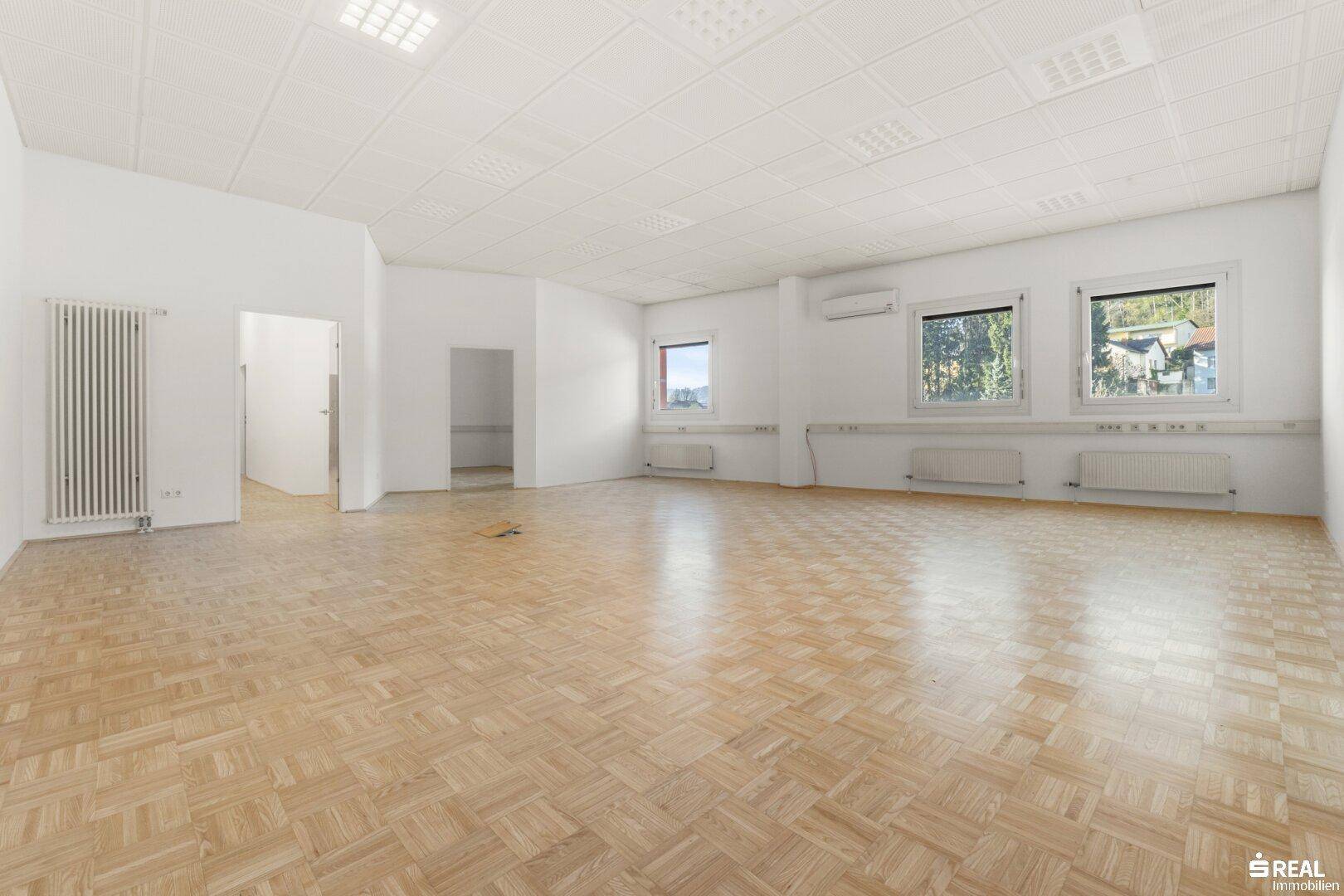 Weitläufiger, heller Büroraum mit Parkettboden, Heizkörpern und Fenstern mit Blick ins Grüne.