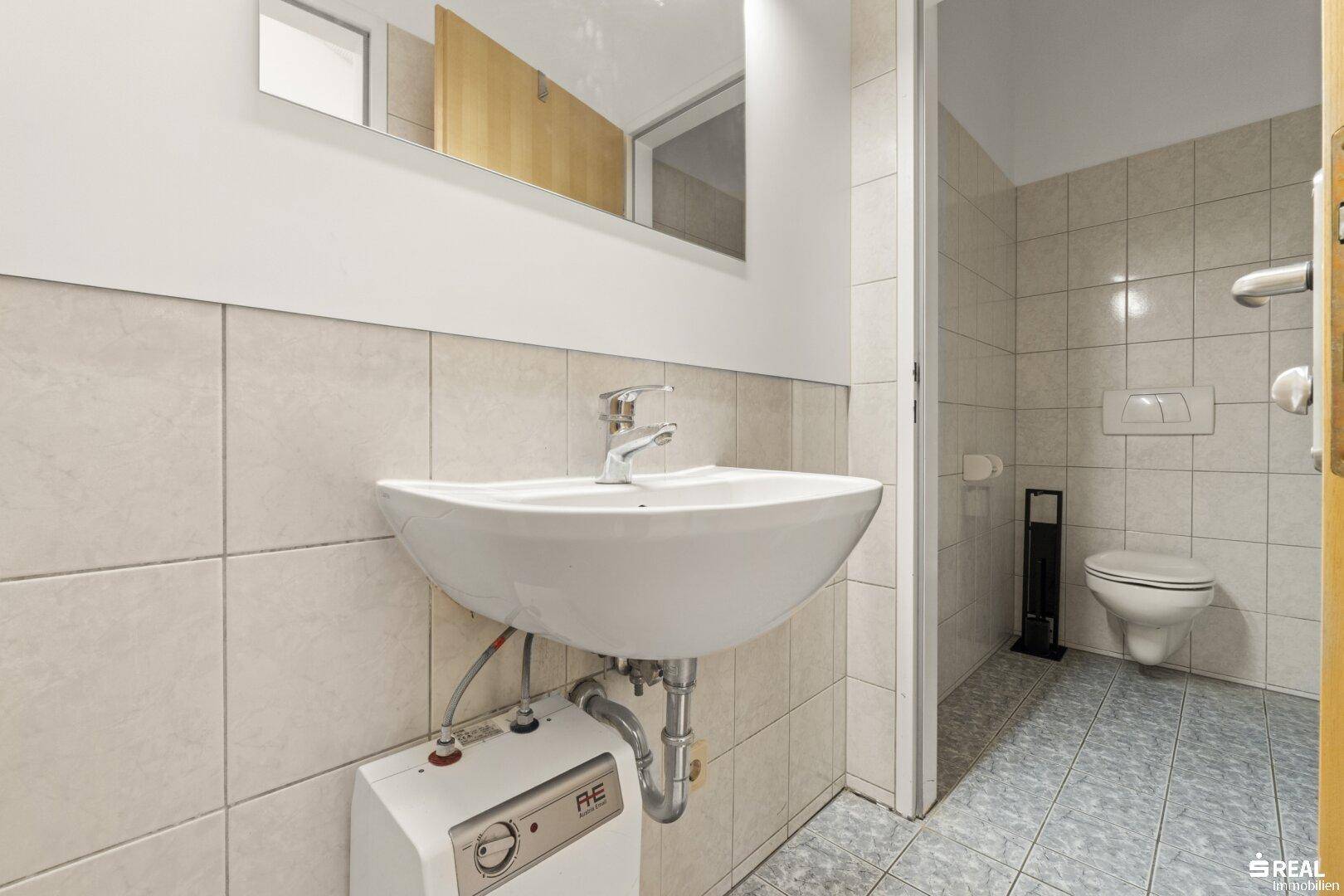 Toilettenraum mit Waschbecken, Spiegel und hellen Fliesen an Wänden und Boden.