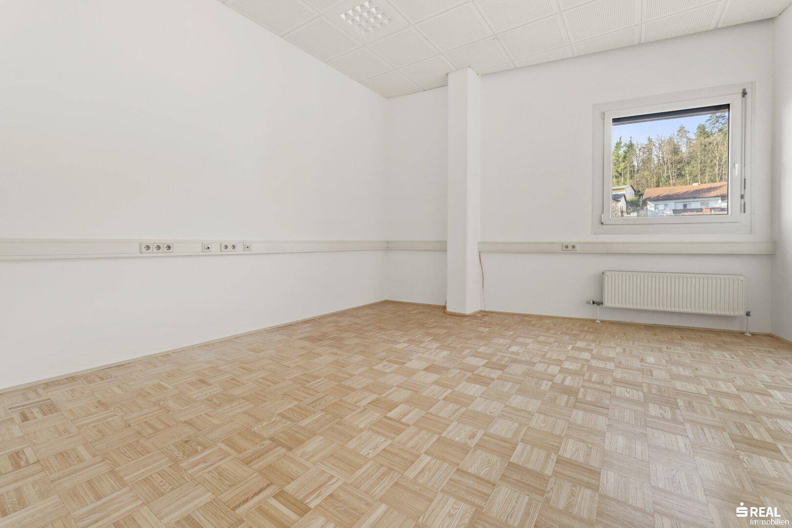 Kleinerer Büroraum mit Parkettboden, weiß gestrichenen Wänden und einem Fenster mit Jalousien.