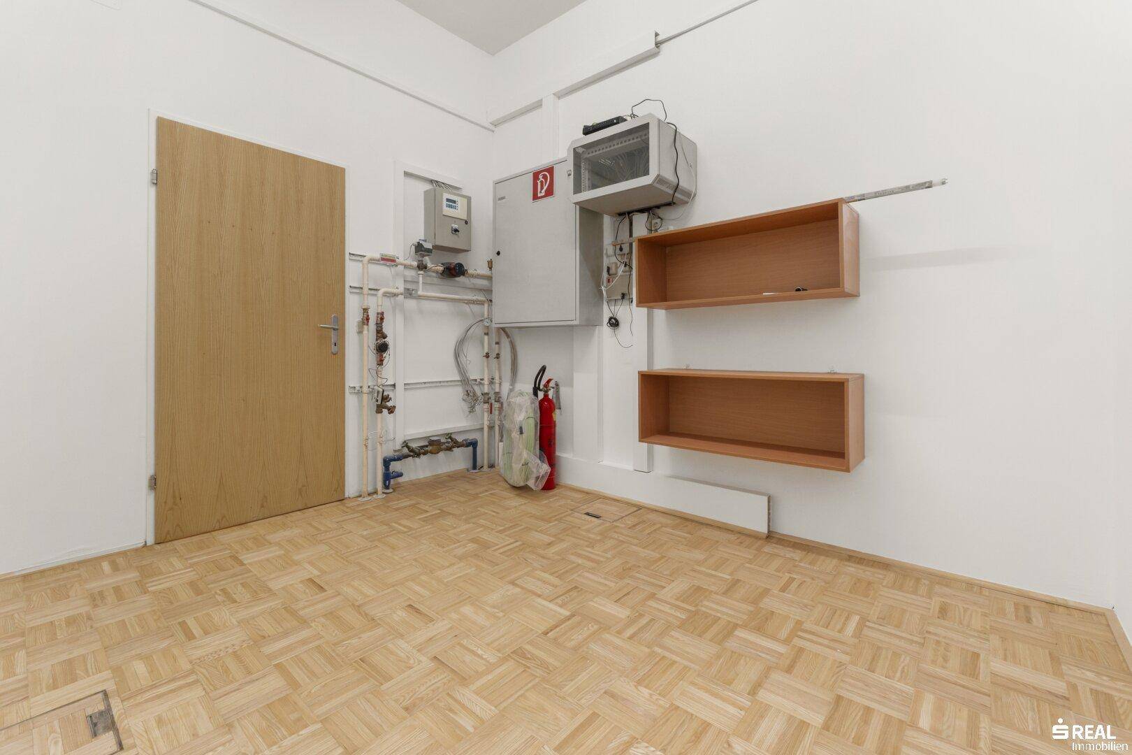 Technikraum mit sichtbaren Installationen, Elektroschaltkasten und einem Feuerlöscher.