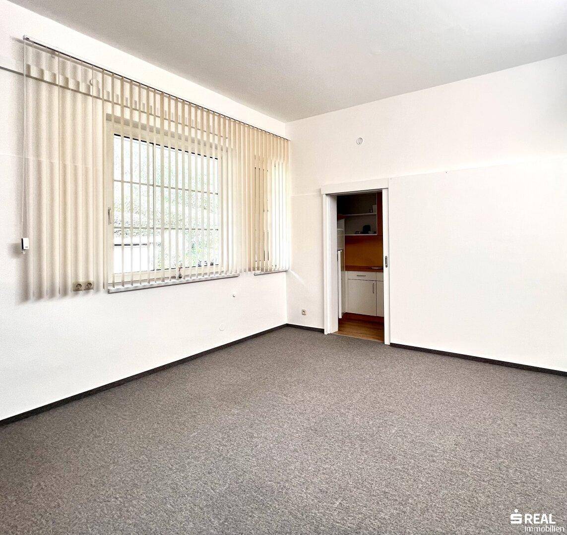 Büroraum mit Teppichboden und Fenster, angrenzend an eine kleine Küchenzeile.