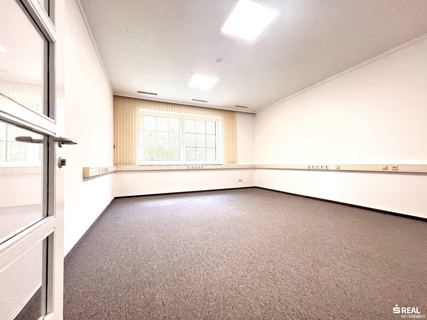 Helles Büro mit Teppichboden, Fenster und Wandleisten für Verkabelung, funktional eingerichtet.