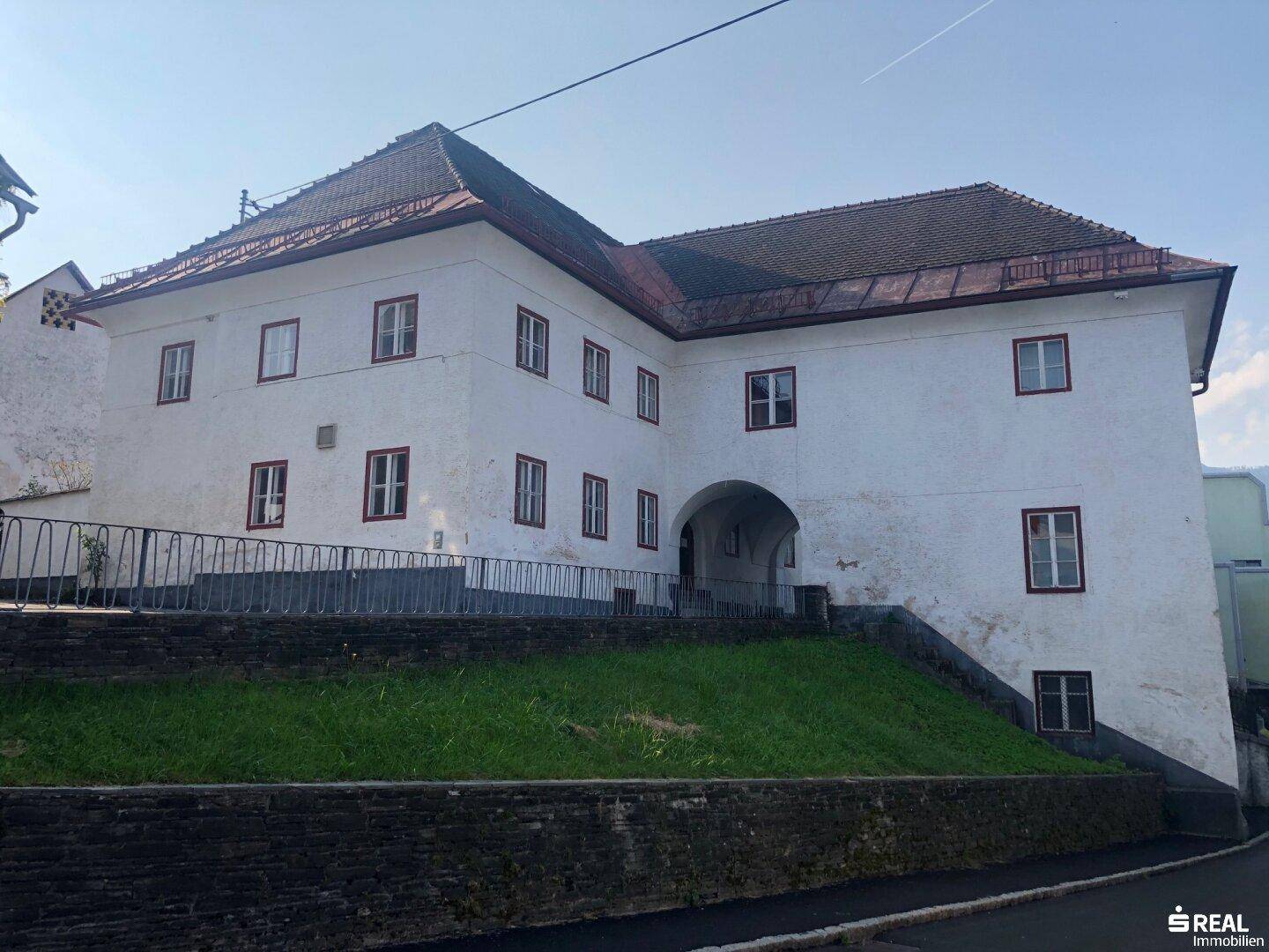 Historisches Gebäude mit weißer Fassade, roten Fensterrahmen und Satteldach unter blauem Himmel.