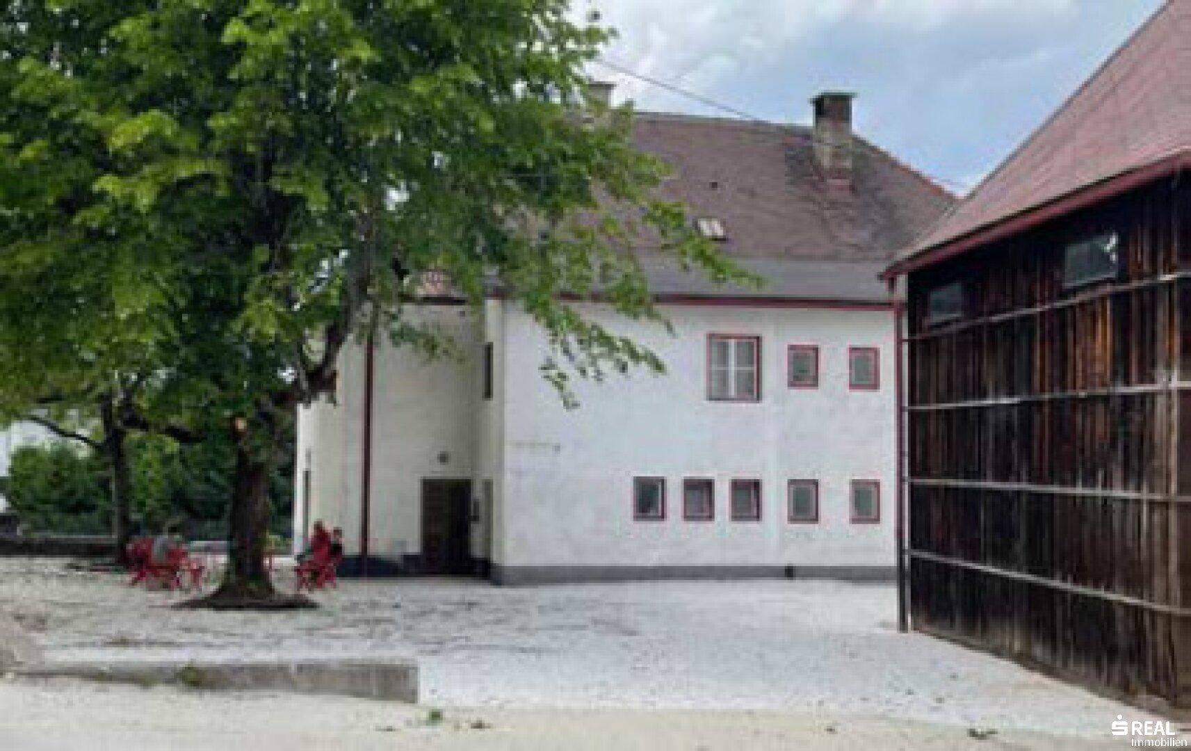 Historisches Gebäude neben einer Scheune aus Holz mit großem Baum auf dem Kiesplatz.