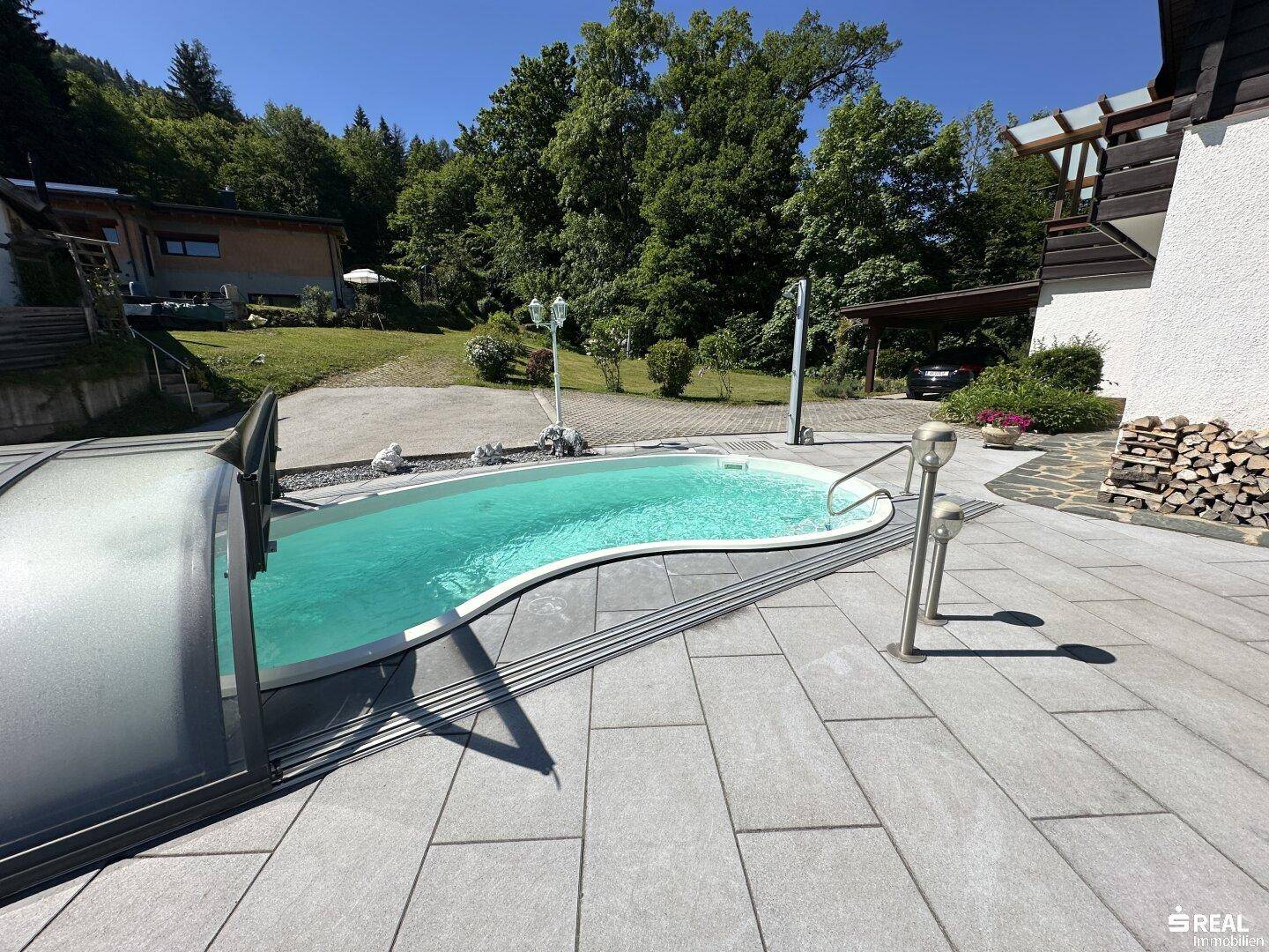 Außenpool mit Abdeckung und gepflasterter Terrasse, umgeben von Bäumen und einem Holzstapel.