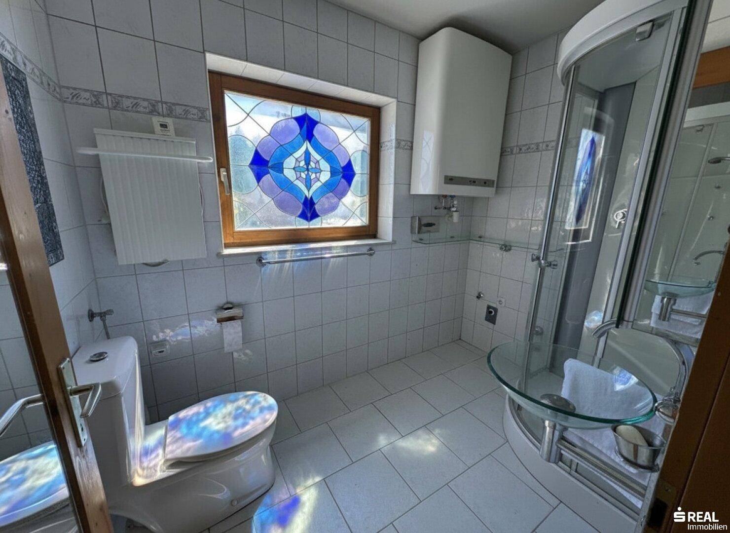 Helles Badezimmer mit Dusche, WC und einem farbigen Buntglasfenster.