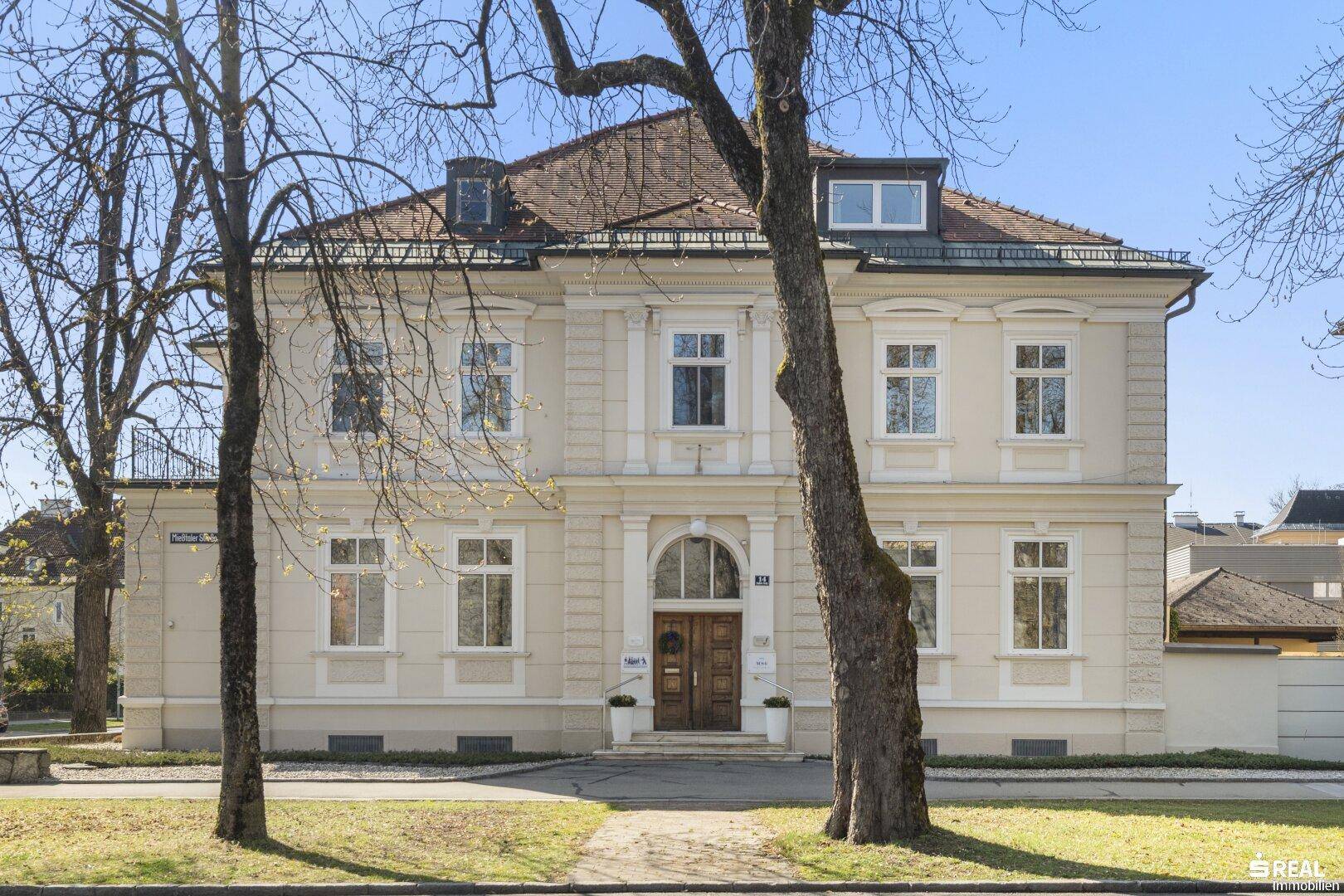 Elegante Fassade eines klassischen Gebäudes mit repräsentativem Eingangsbereich und hohen Bäumen.