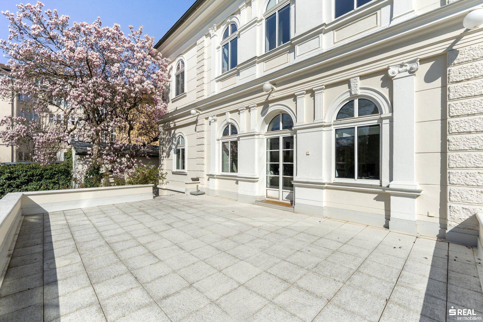 Geräumige, gepflasterte Terrasse mit Blick auf blühende Bäume und die klassische Fassade.