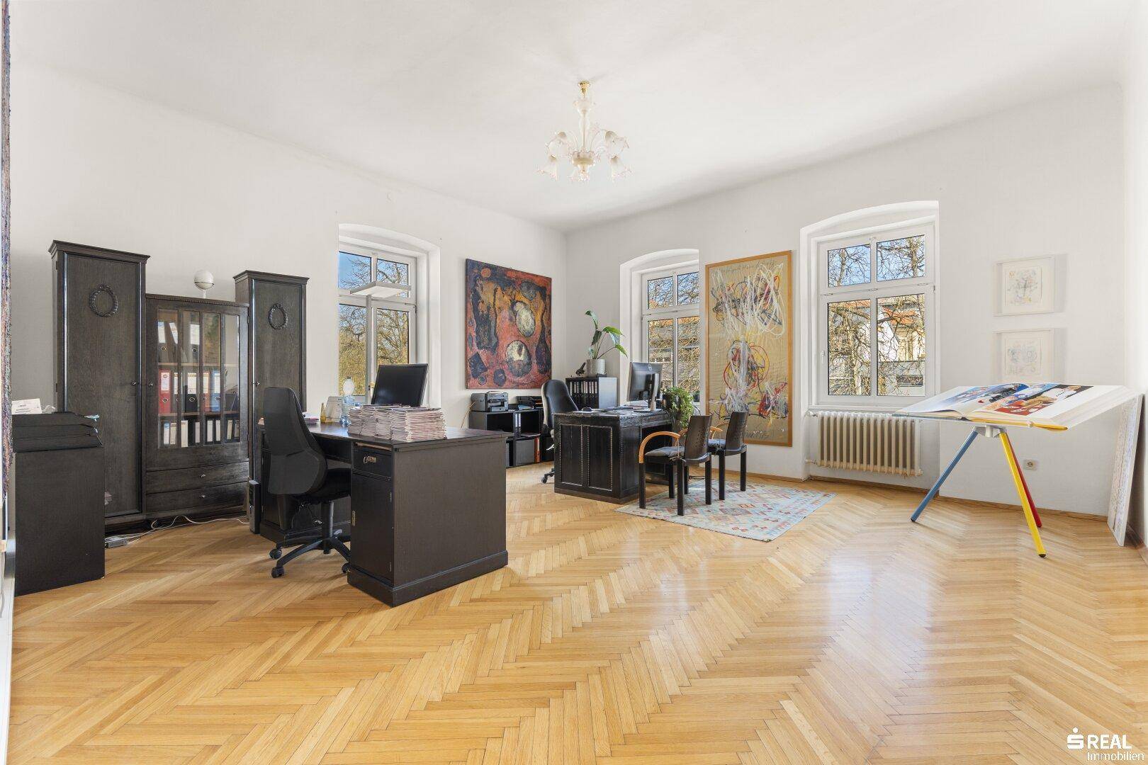 Helles Büro mit Fischgrätparkett, dunklen Holzmöbeln und Blick ins Grüne durch die Fenster.