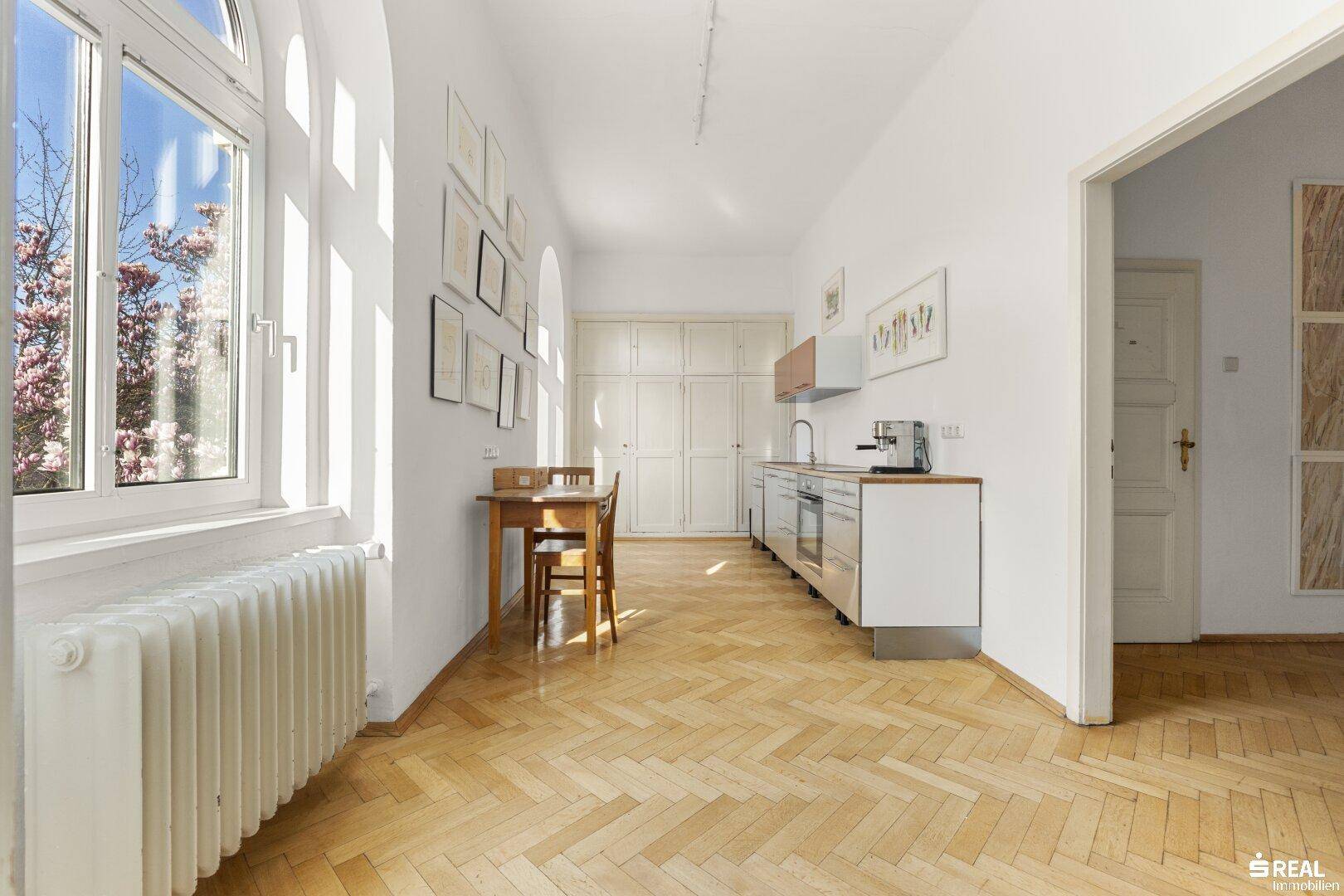 Schmale Küche mit Holzboden, Einbauschränken und einem kleinen Essbereich am Fenster.