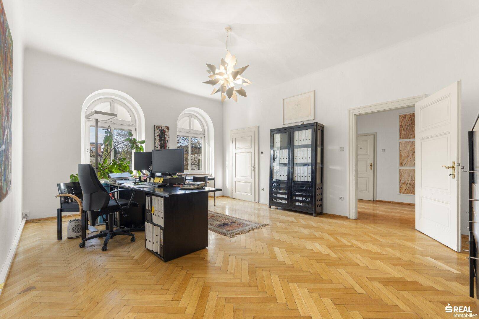 Großes Büro mit Fischgrätparkett, hohen Fenstern und einem modernen Schreibtischbereich.