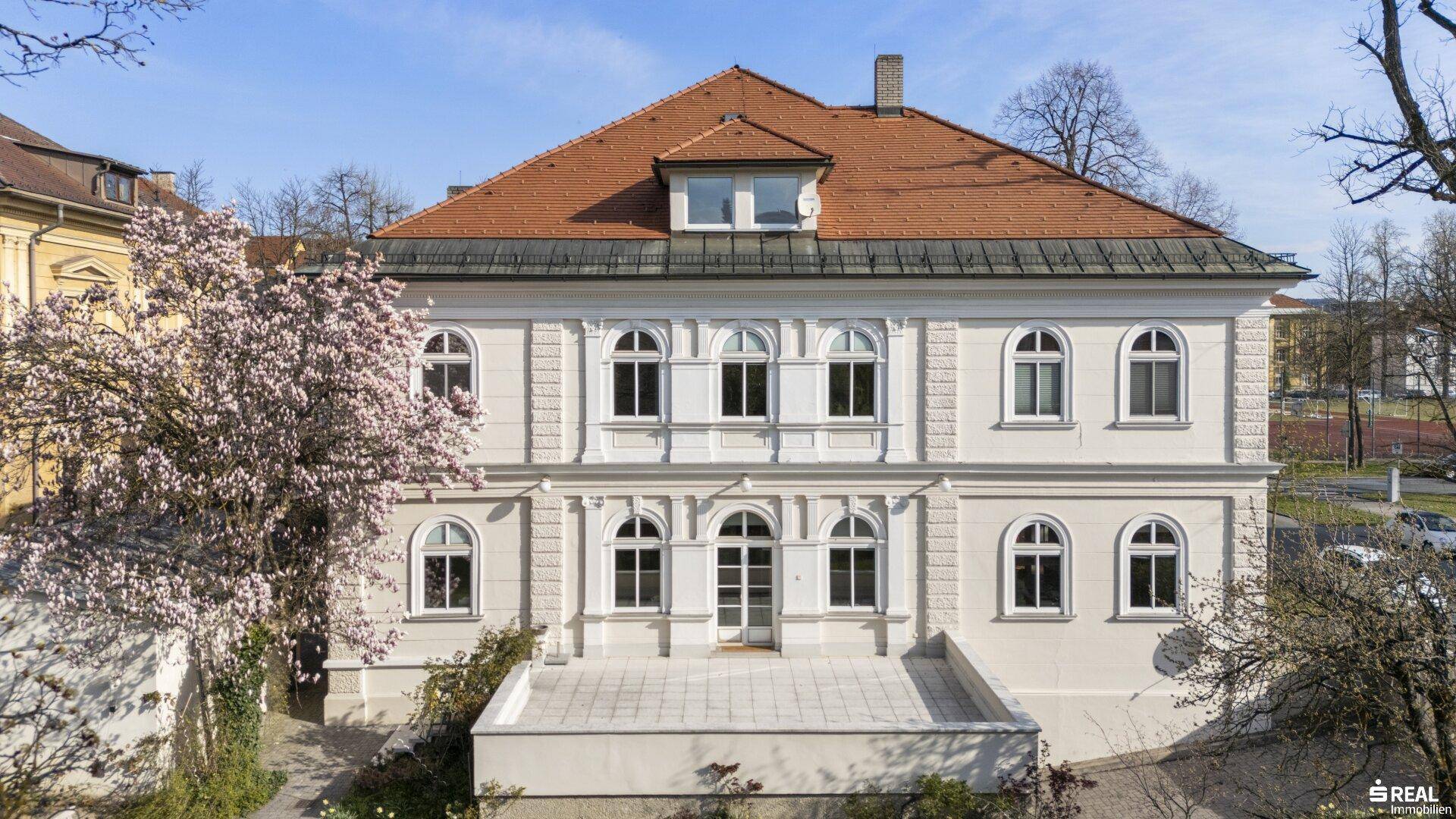 Rückseite des Gebäudes mit großer, gepflasterter Terrasse und Blick auf die Umgebung.
