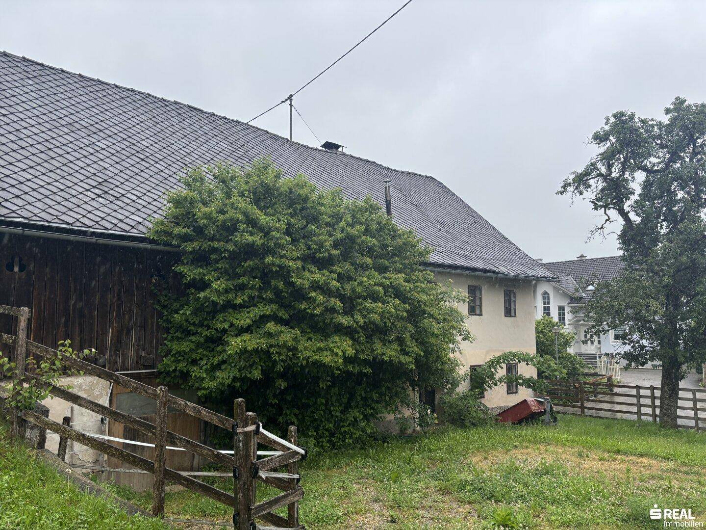 Ansicht des historischen Gebäudes mit großem Gartenbereich und üppiger Vegetation an einem regnerischen Tag.