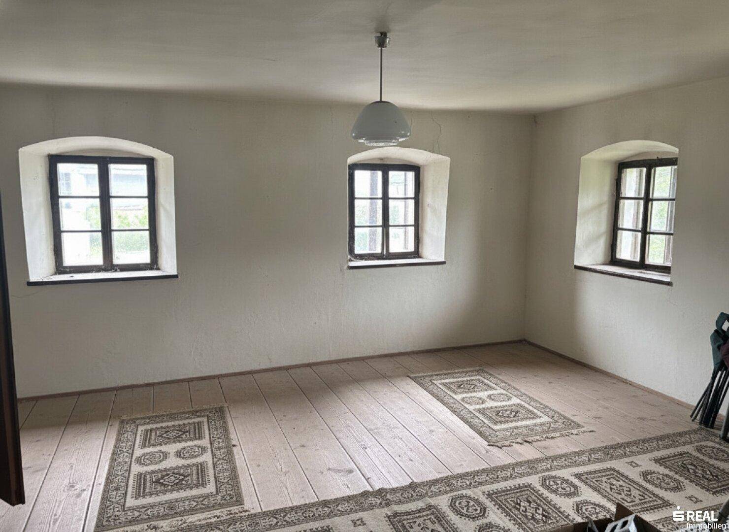 Geräumiges Zimmer mit Holzdielenboden, traditionellen Teppichen und drei Fenstern für viel Licht.
