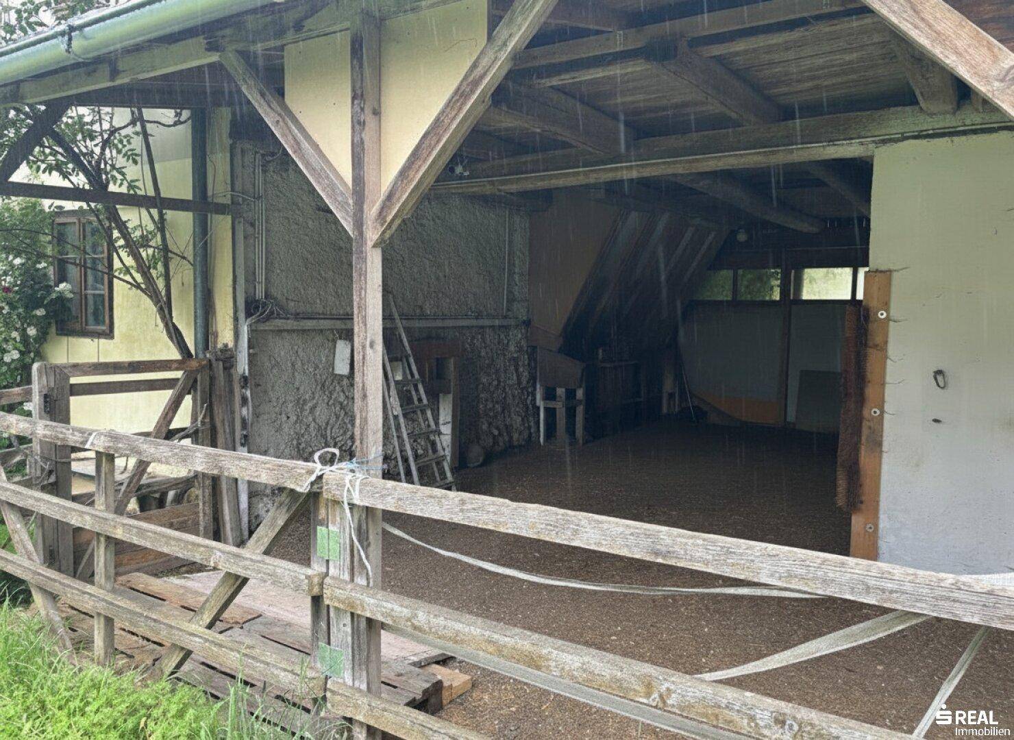 Überdachter Außenbereich mit Holzbalken und unbefestigtem Boden, möglicherweise ein Stall oder Lager.