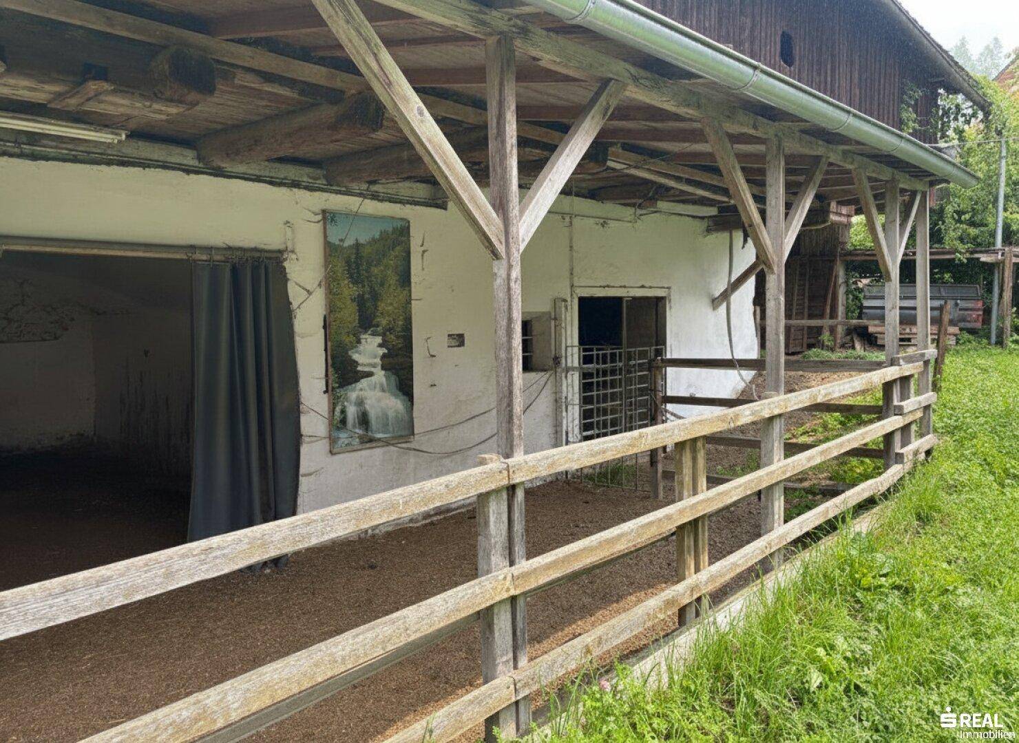 Länglicher überdachter Bereich mit Holzzaun und Blick auf eine ländliche Umgebung.
