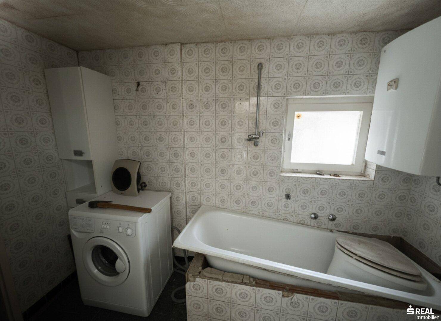 Badezimmer mit alten Fliesen, Badewanne und Waschmaschine, renovierungsbedürftig.