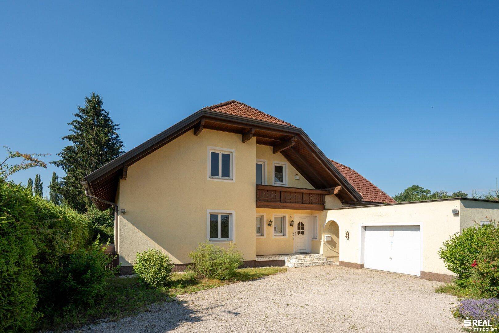 Vorderansicht des Hauses mit gelber Fassade, Holzbalkon und gepflegter Auffahrt unter blauem Himmel.