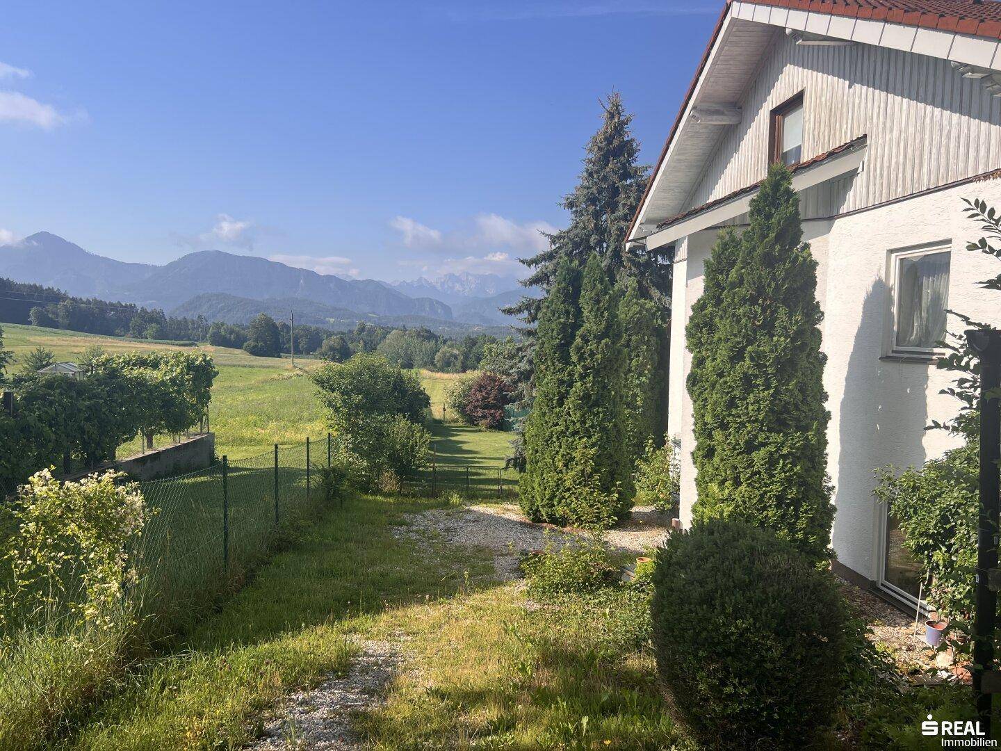 Seitenansicht eines weißen Hauses mit rotem Dach, umgeben von hohen Bäumen und einem weiten Feld mit Bergblick.