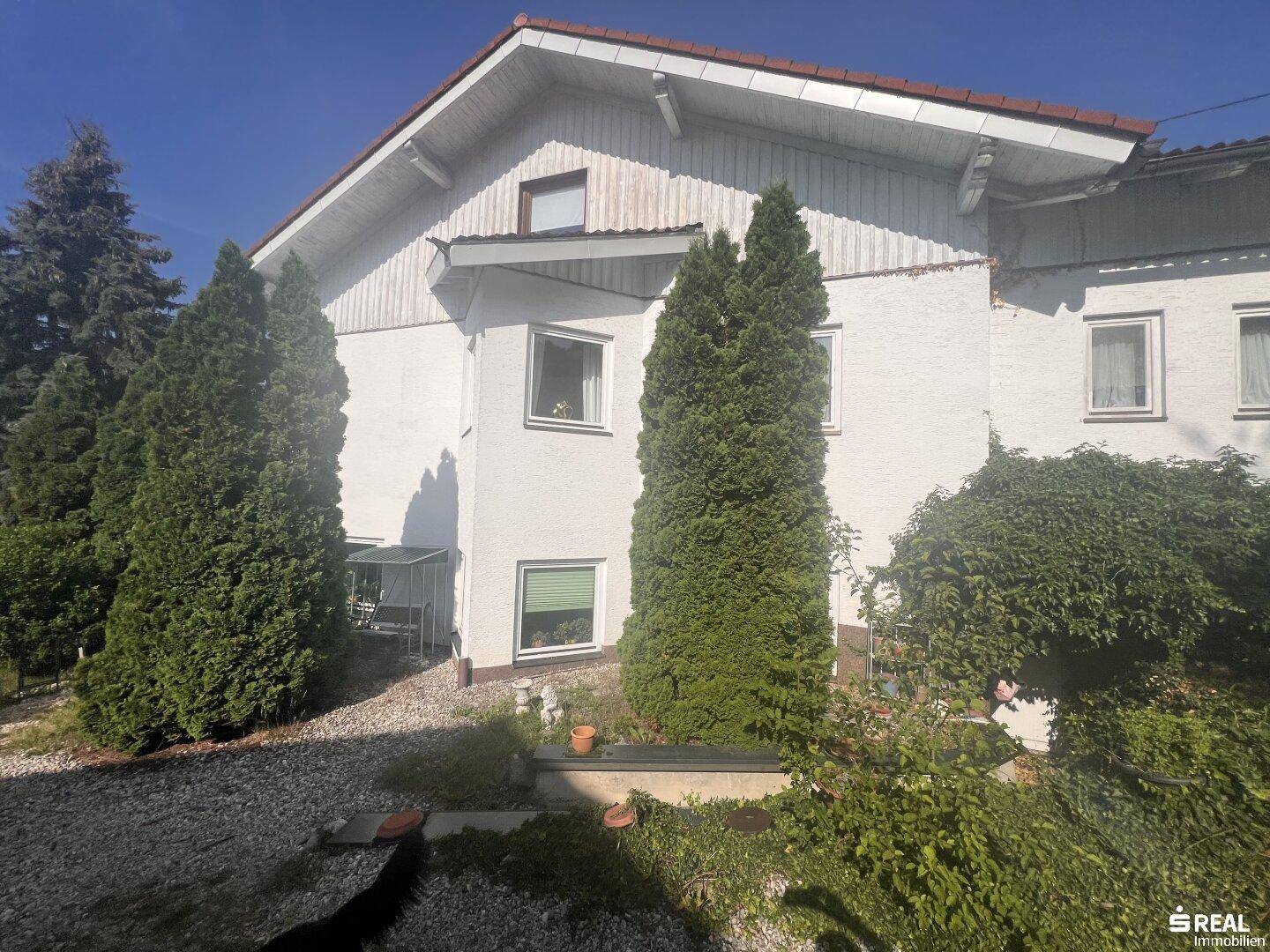 Ansicht der Hausfassade mit weißem Putz und rotem Dach, umgeben von hohen Nadelbäumen und einem Kiesweg.