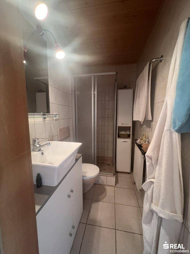 Funktionales Badezimmer mit weißen Fliesen, Duschkabine, Waschbecken und WC, ergänzt durch eine Holzdecke.