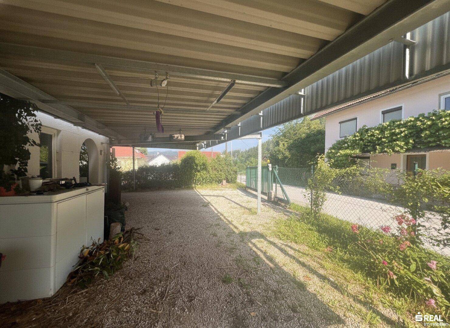 Geräumiger Carport-Bereich mit Kiesboden, der zu einem grünen Tor führt, umgeben von Hecken und Bäumen.