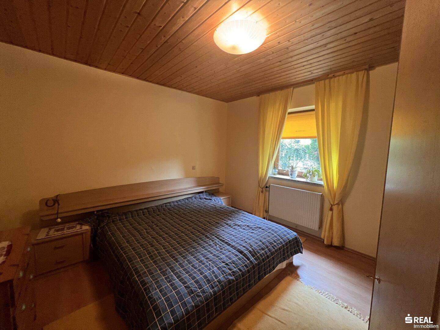 Helles Schlafzimmer mit Doppelbett, gelben Vorhängen und einer warmen Holzdecke, auf Holzboden.