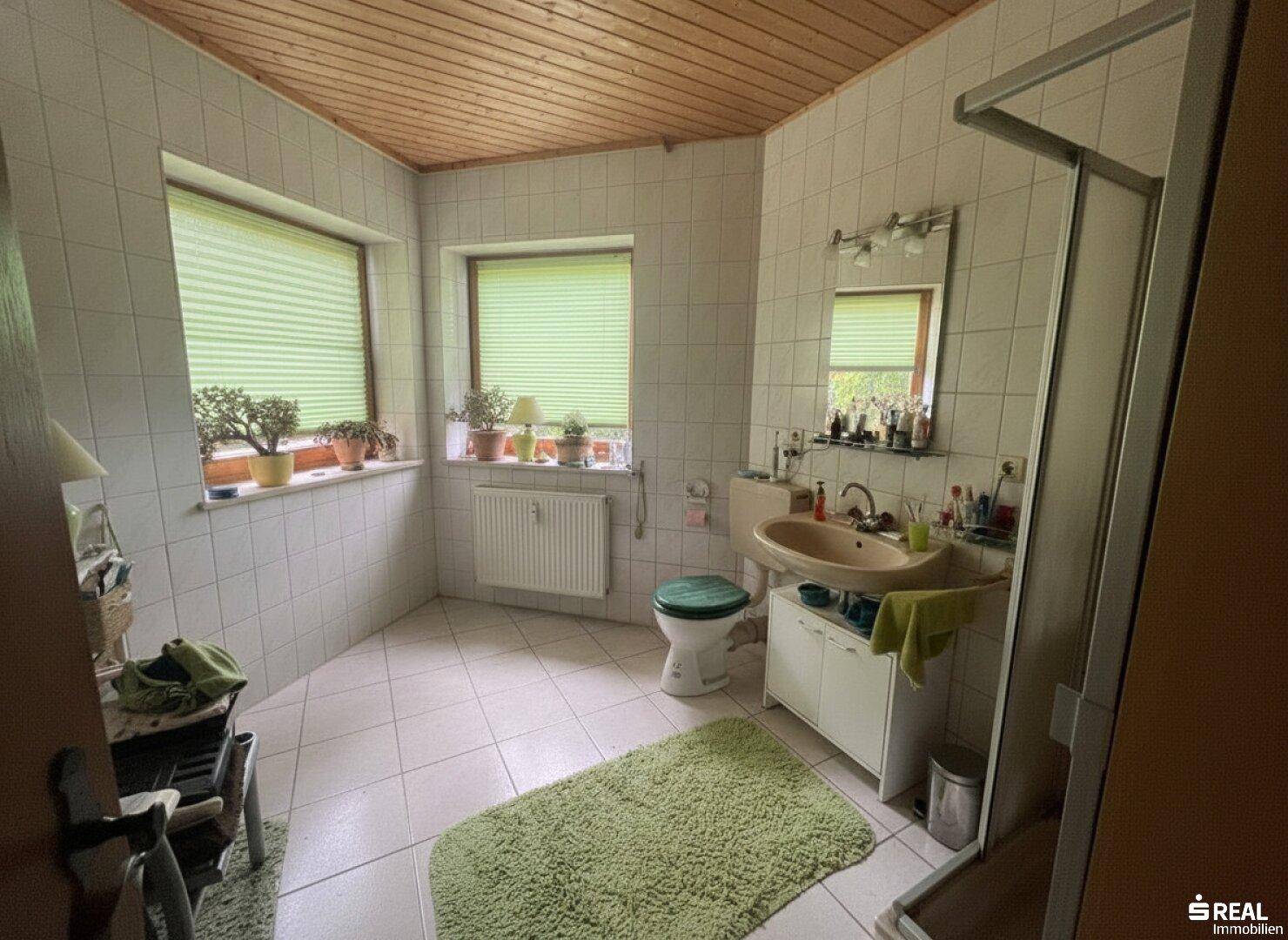 Geräumiges Badezimmer mit weißen Fliesen, Duschkabine und zwei Fenstern, ausgestattet mit einer Holzdecke.
