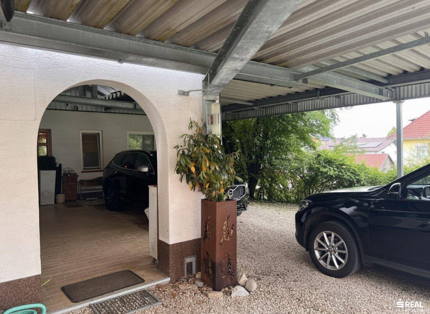 Eingangsbereich zum Carport mit einem Bogen, der zu einem geschützten Parkbereich auf Kiesboden führt.