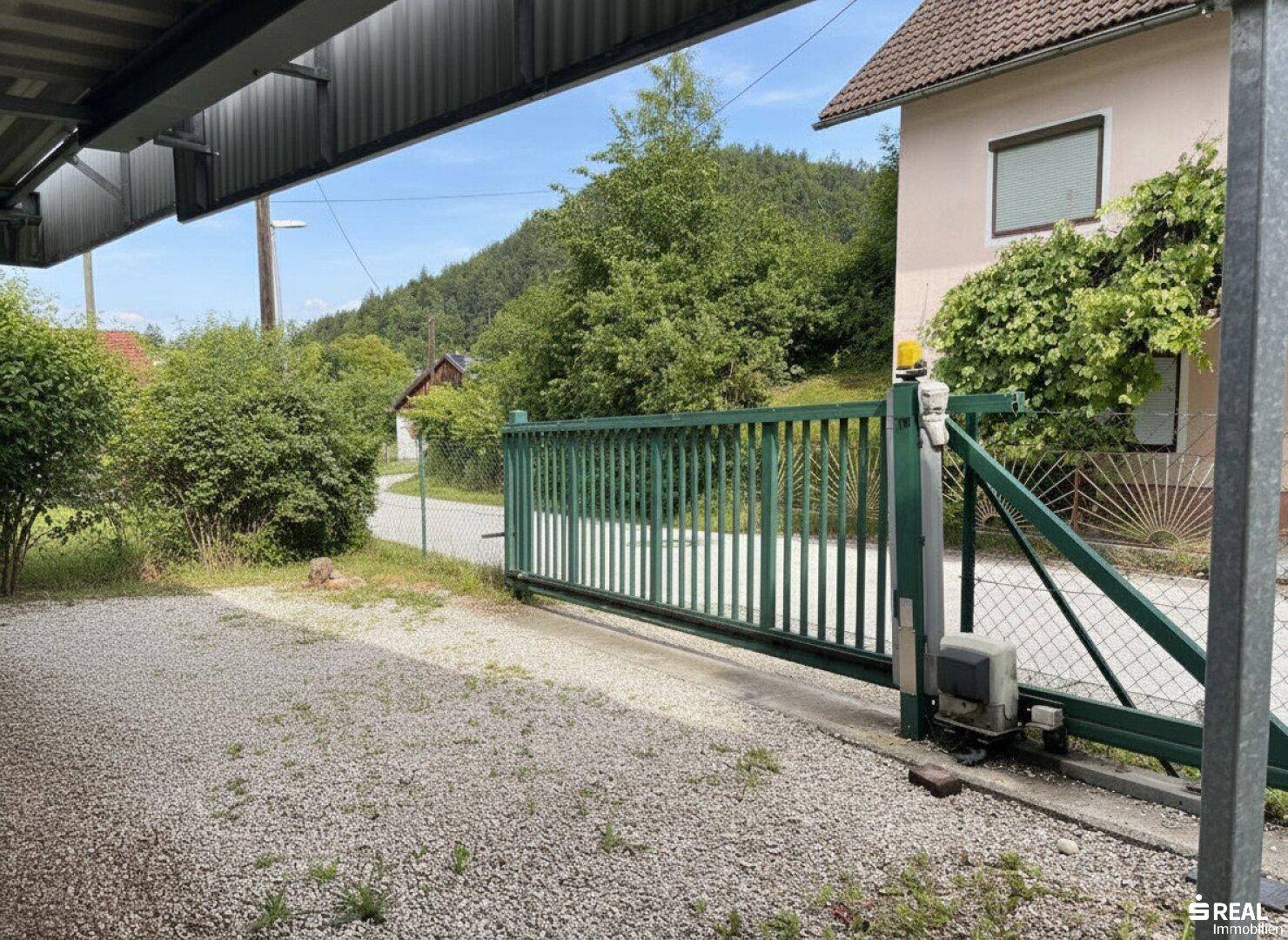 Grünes Metalltor an einer Kiesauffahrt, bietet Zugang zum Grundstück mit Blick auf die umliegenden Hügel.