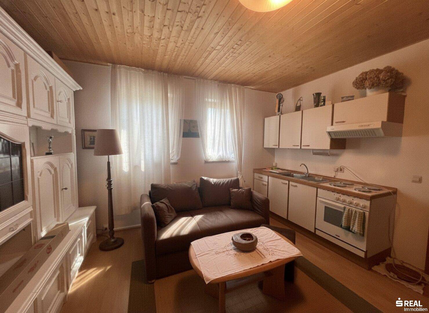 Kombinierter Wohnbereich mit Sofa und kleiner Küchenzeile, weißen Schränken und Holzdecke, auf Holzboden.