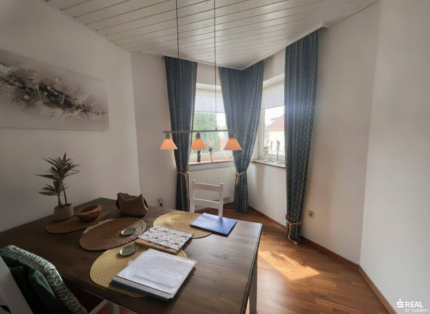 Heller Essbereich mit Holztisch und Stühlen, ausgestattet mit einem Eckfenster und dunklen Vorhängen.