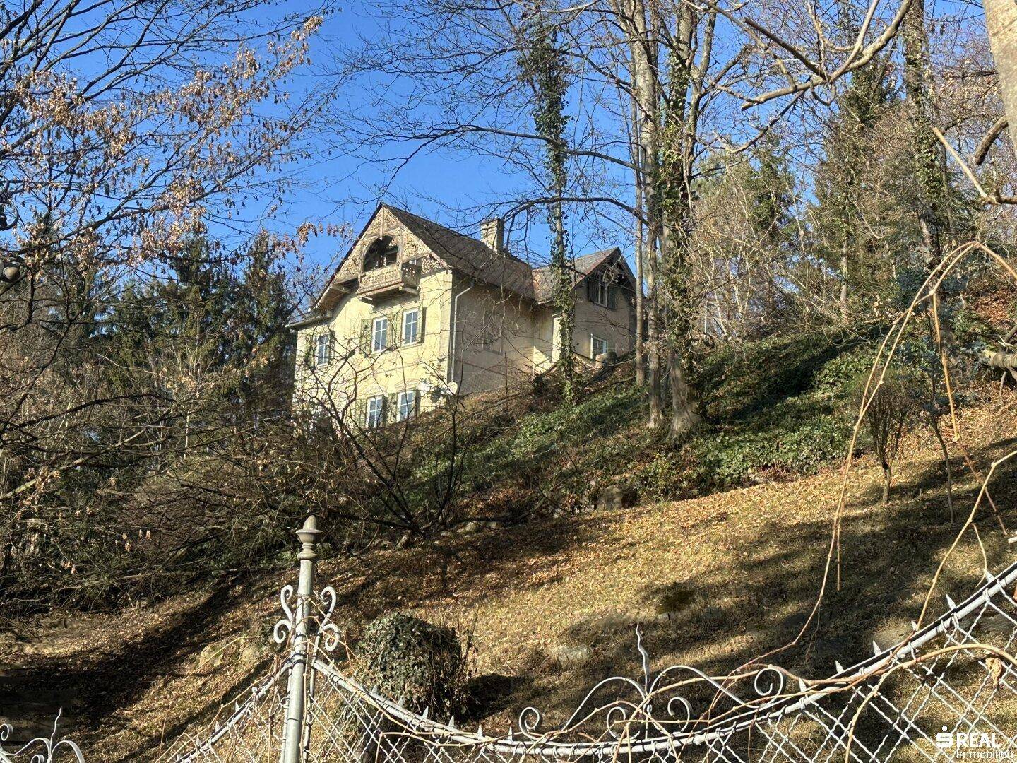 Historisches Haus auf einem Hügel, umgeben von kahlen Bäumen im Winter, mit einem dekorativen Metallzaun im Vordergrund.