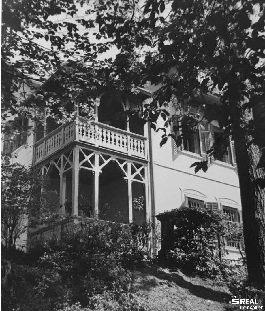 Schwarz-Weiß-Aufnahme eines historischen Hauses mit einer großen, verzierten Holzveranda, umgeben von Bäumen.