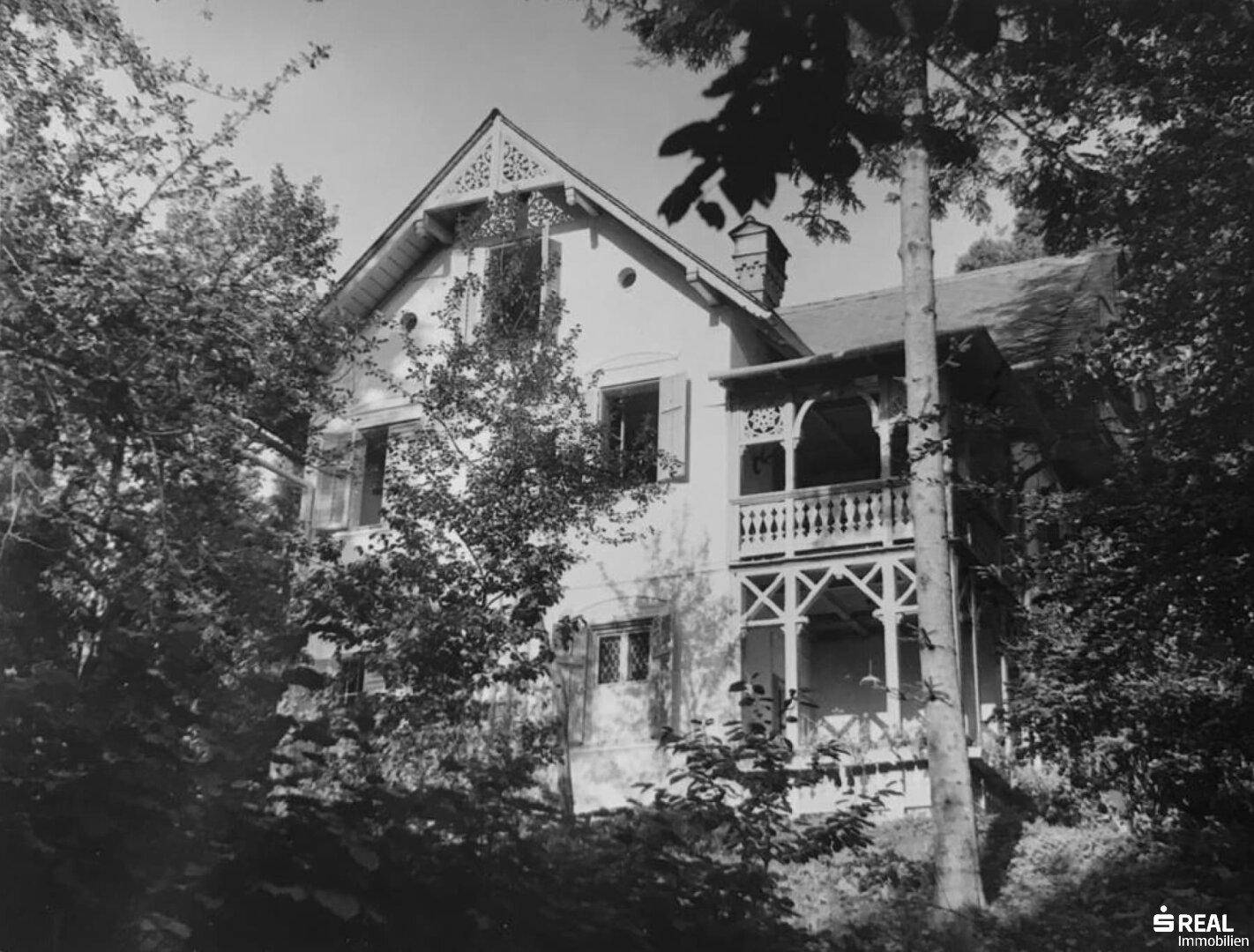 Schwarz-Weiß-Aufnahme des historischen Hauses mit kunstvollem Balkon, umgeben von dichtem Baumbestand.