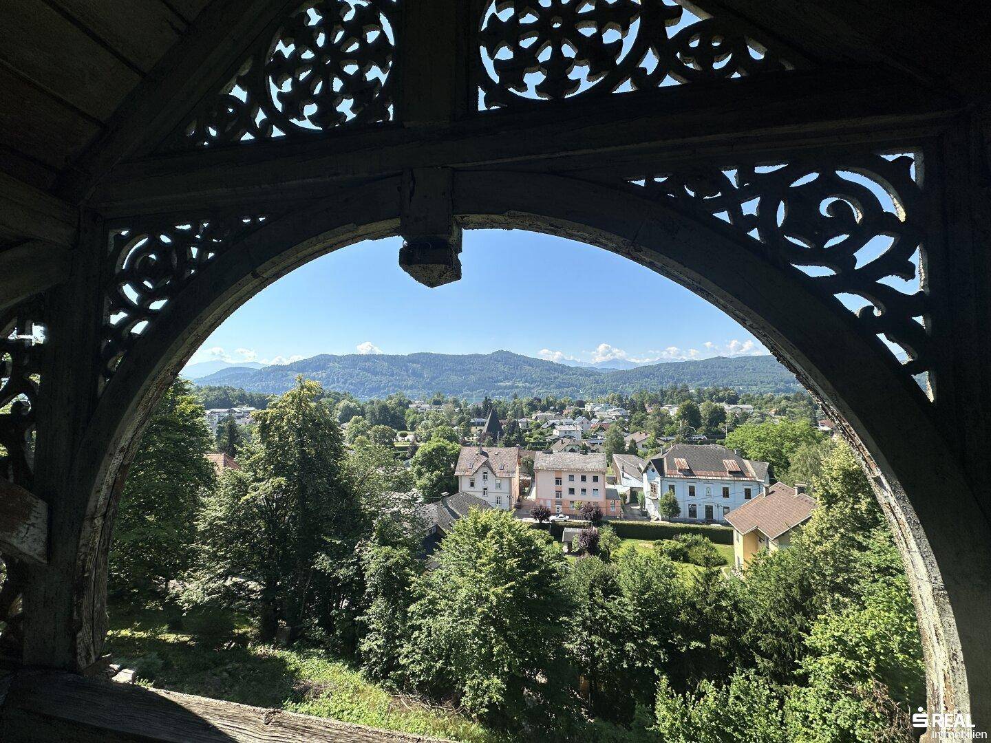 Blick von einem kunstvollen Holzbalkon mit üppiger grüner Vegetation und Bergblick an einem klaren Sommertag.