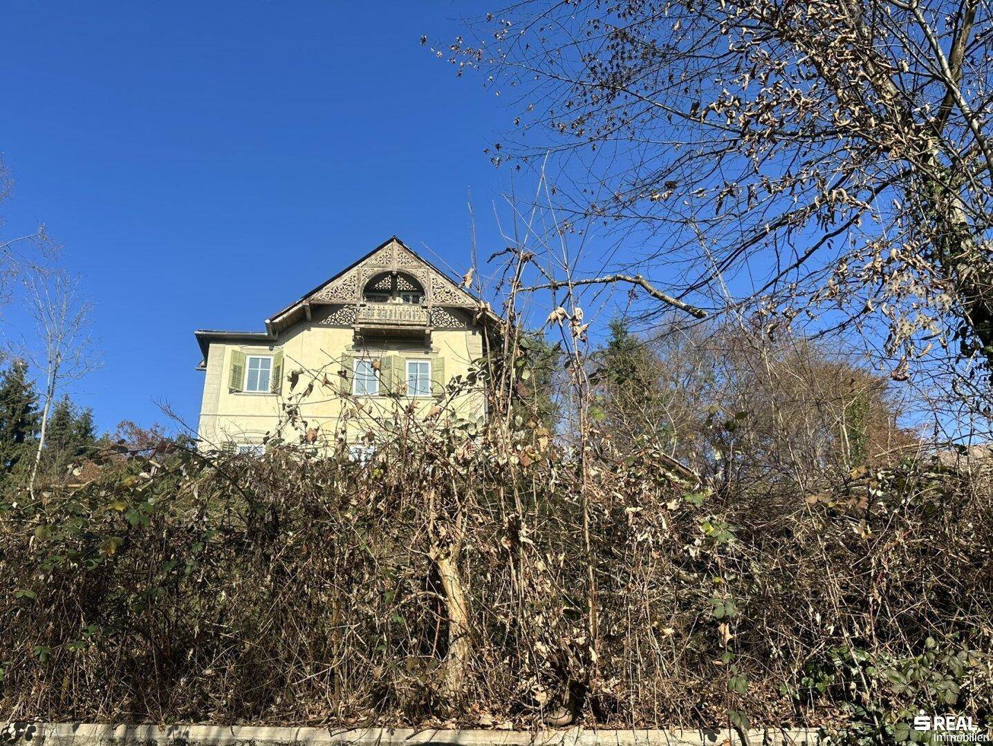 Das historische Haus ist hinter dichtem Bewuchs und Bäumen sichtbar, mit einem klaren blauen Himmel darüber.