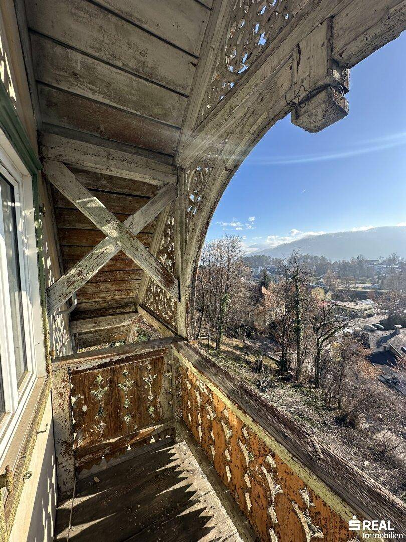 Detailansicht eines verwitterten, kunstvoll geschnitzten Holzbalkons mit Blick auf die Umgebung und sonnigem Himmel.